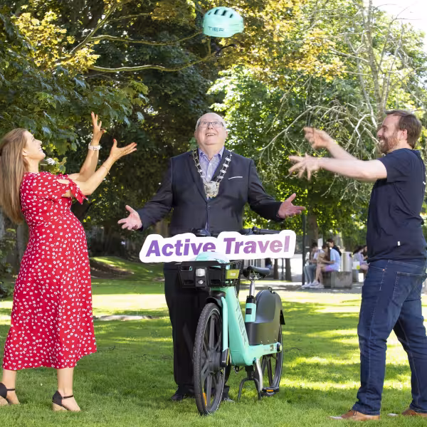 Woman in red dres throws bike helmet to man in navy t-shirt while Mayor stands between them holding e-bike