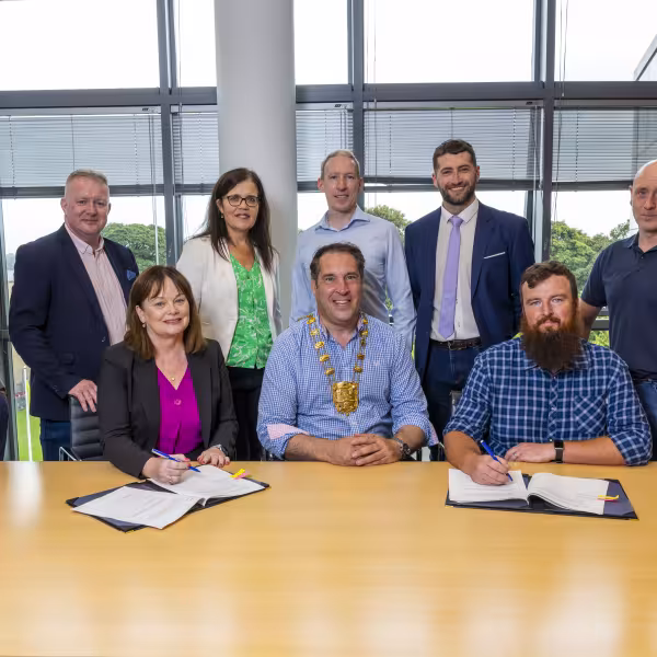 Mayor wearing chain of office signs paper contract with man on right seated and woman on left seated. Team of men and women standing at rear.