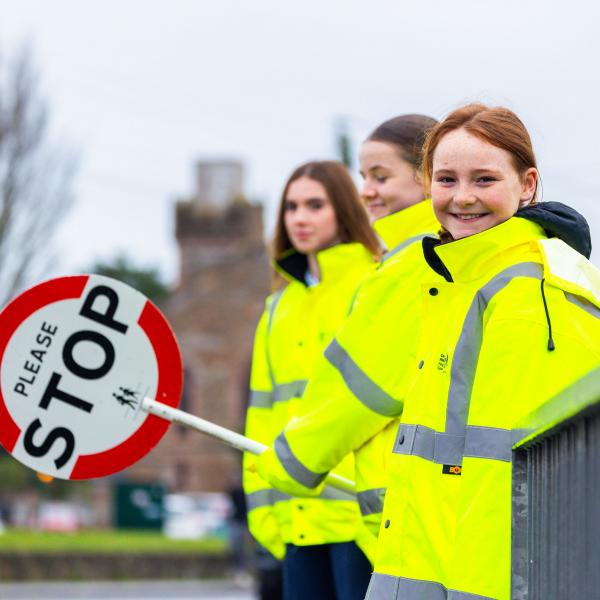 3 girls in yellow hi-vis jackets standing facing a road viewed from side. One is holding a stop sign.