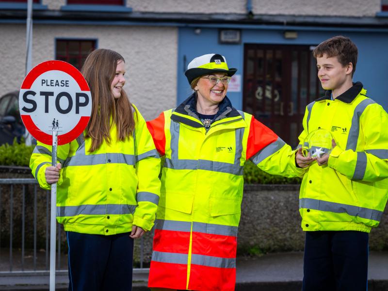School warden wearing yellow and orange hi-vis uniform and holding stop sign stands with two students in hi-vis jackets