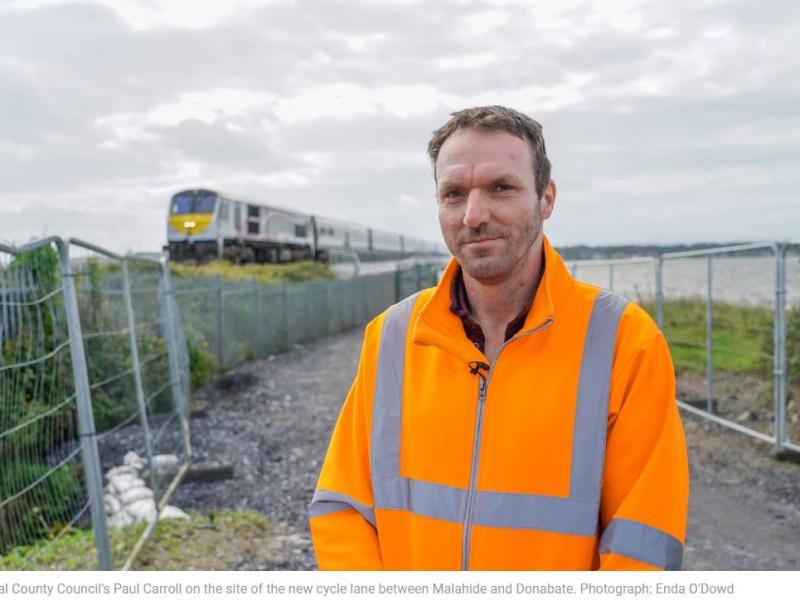 Man in orange jacket stands on site with waterside and train in background