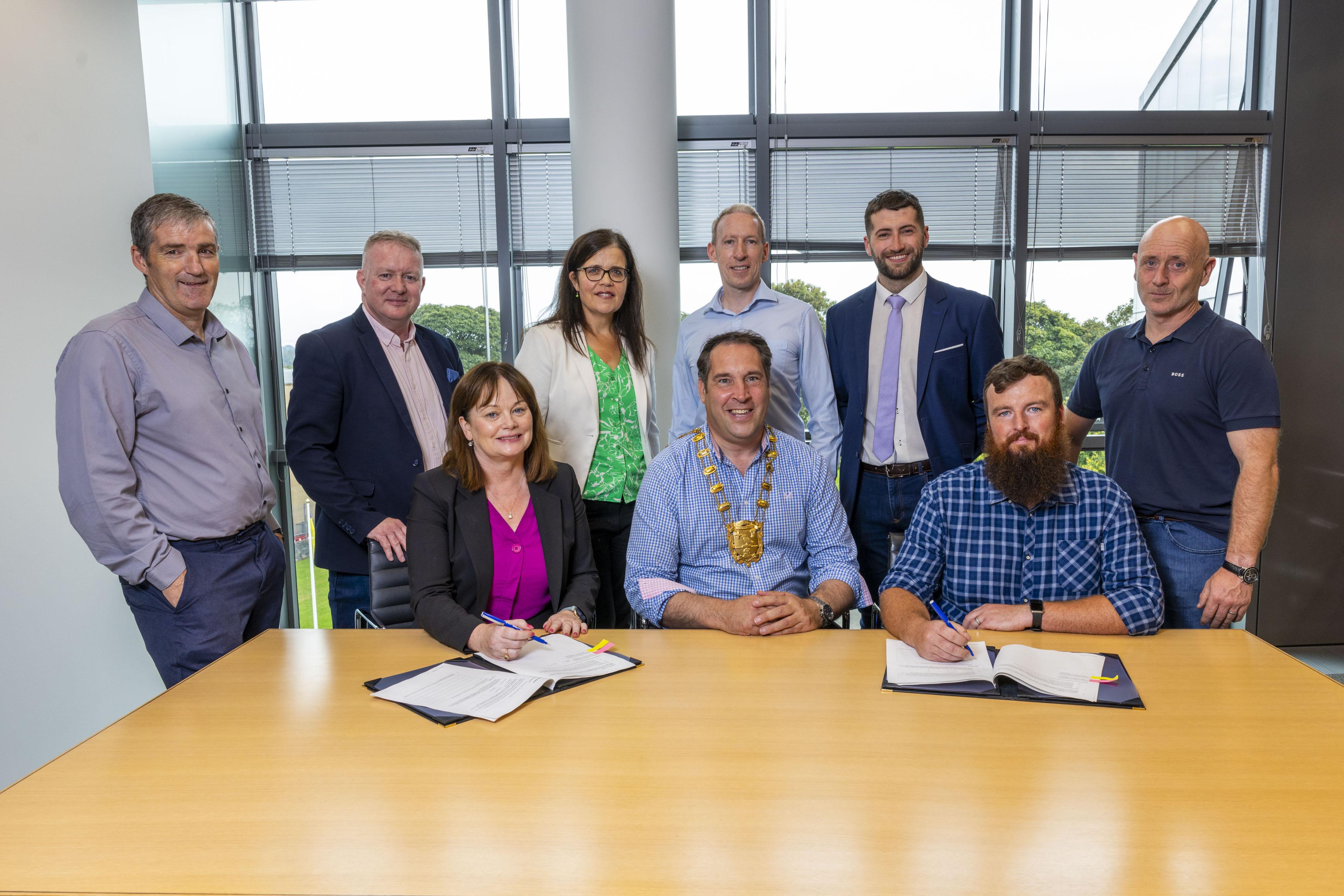 Mayor wearing chain of office signs paper contract with man on right seated and woman on left seated. Team of men and women standing at rear.