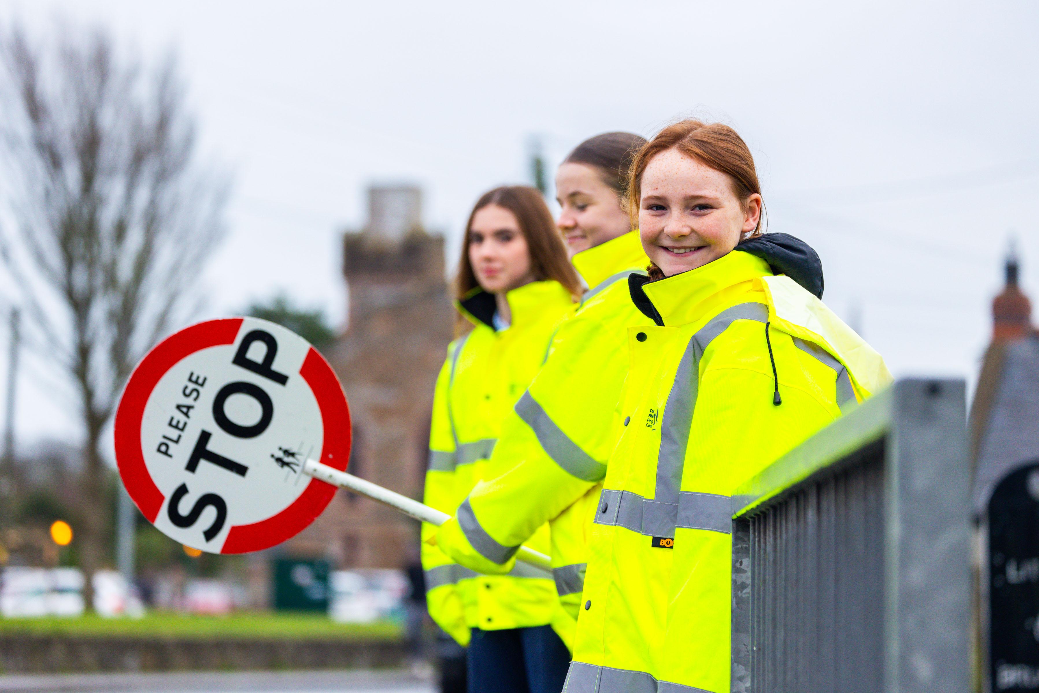 3 girls in yellow hi-vis jackets standing facing a road viewed from side. One is holding a stop sign.